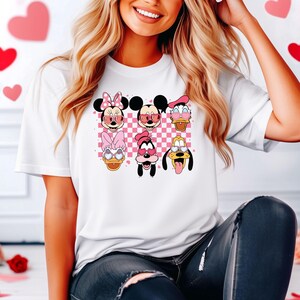 May include: White t-shirt with a pink and white checkered background featuring cartoon characters from Disney's Mickey Mouse franchise wearing heart-shaped sunglasses. The characters include Mickey Mouse, Minnie Mouse, Donald Duck, Goofy, and Pluto.
