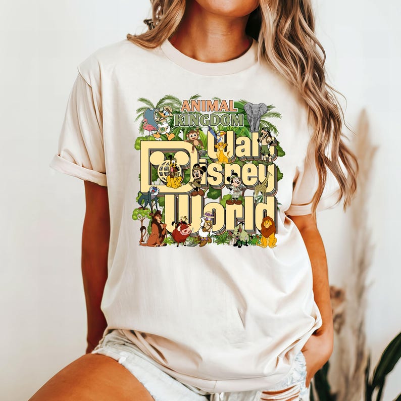 Pu&ograve; includere: Una maglietta beige con una stampa grafica del logo di Walt Disney World e vari personaggi animali del parco Animal Kingdom. Il testo "Animal Kingdom" &egrave; sopra il logo e il testo "Walt Disney World" &egrave; sotto il logo.