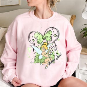 May include: Pink sweatshirt with a green and black graphic of Tinker Bell with a flower, a castle, and a bow. The text "Tinker Bell" and "Bell" are written on the graphic.