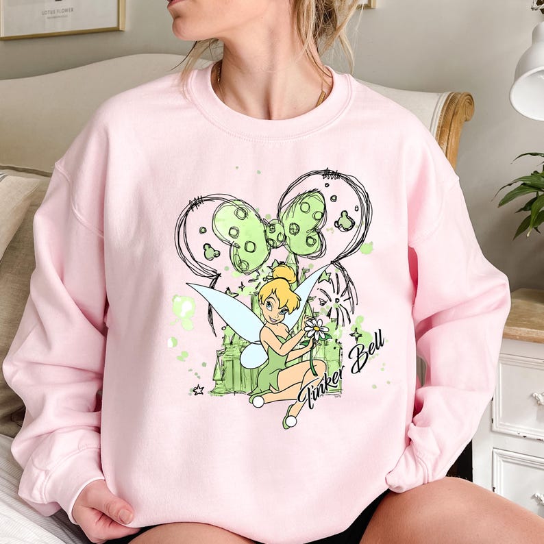May include: Pink sweatshirt with a green and black graphic of Tinker Bell with a flower, a castle, and a bow. The text "Tinker Bell" and "Bell" are written on the graphic.