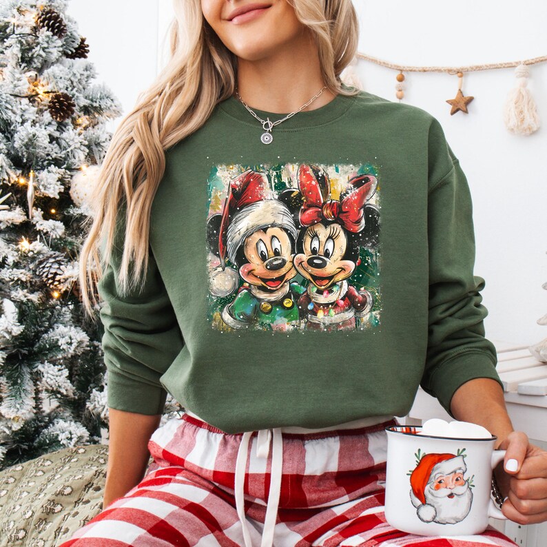 May include: Green sweatshirt featuring a festive graphic of Mickey and Minnie Mouse in Christmas attire. The image shows Mickey wearing a Santa hat and Minnie with a red bow. The sweatshirt is paired with red and white plaid pajama pants.
