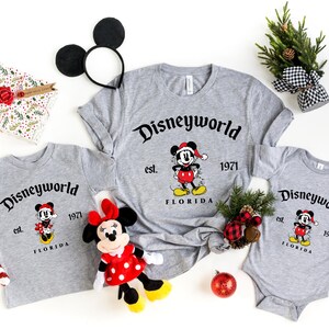May include: Three gray t-shirts with a Mickey Mouse design and the text "Disneyworld est. 1971 Florida". The shirts are in different sizes, with the largest shirt having a long sleeve. The shirts are surrounded by Christmas decorations, including a candy cane, a red ornament, and a small Christmas tree.