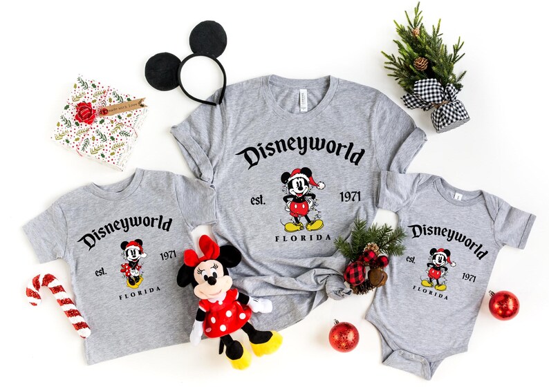 May include: Three gray t-shirts with a Mickey Mouse design and the text "Disneyworld est. 1971 Florida". The shirts are in different sizes, with the largest shirt having a long sleeve. The shirts are surrounded by Christmas decorations, including a candy cane, a red ornament, and a small Christmas tree.