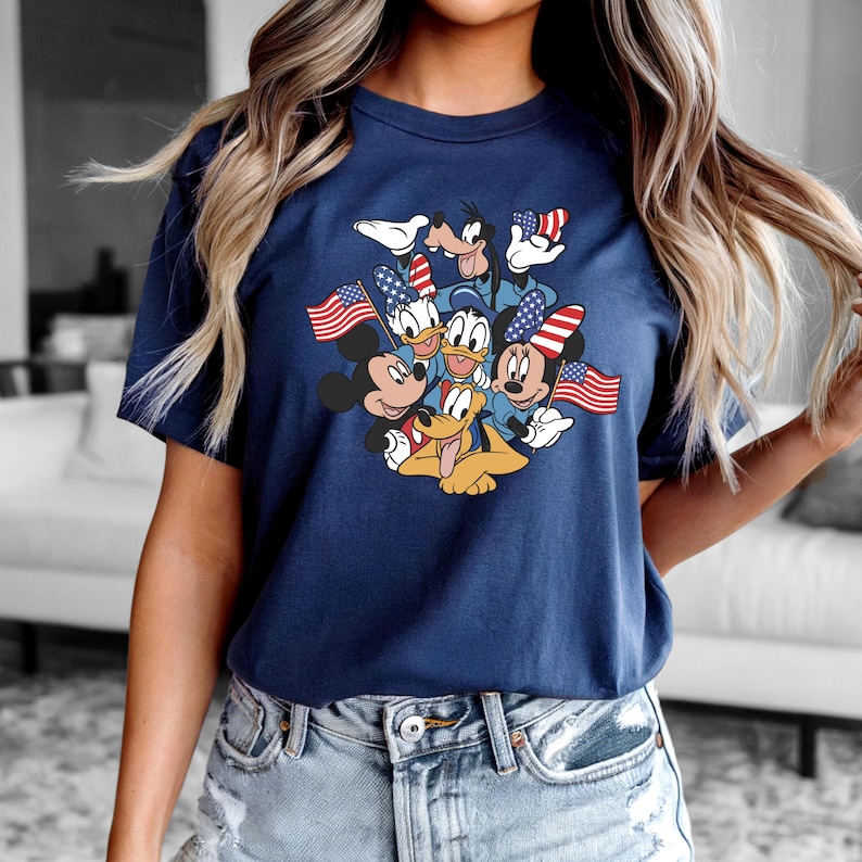 May include: Navy blue t-shirt featuring a graphic of Mickey Mouse, Minnie Mouse, Donald Duck, Daisy Duck, Goofy, and Pluto holding American flags. The characters are in patriotic attire. The shirt is short-sleeved.