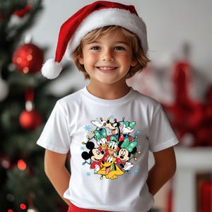 K&ouml;nnte beinhalten: Ein Kind tr&auml;gt ein wei&szlig;es T-Shirt mit einer bunten Grafik von Disney-Figuren, darunter Micky und Minnie Maus, Goofy und Pluto, umgeben von Weihnachtslichtern und Schneeflocken. Das Kind tr&auml;gt auch einen roten Weihnachtsmannhut.