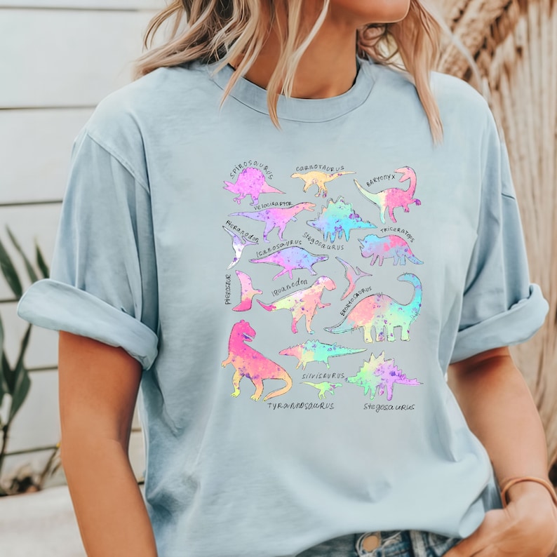 May include: A light blue t-shirt with a colorful graphic print of various dinosaurs. The dinosaurs are outlined in black and filled with a rainbow gradient. The names of each dinosaur are written in black text above each dinosaur.