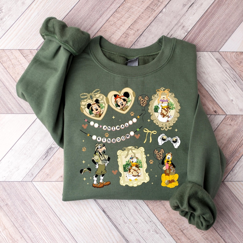 May include: An olive green sweatshirt featuring various Disney characters in decorative frames. The design includes Mickey and Minnie Mouse in heart-shaped frames, along with the text "ANIMAL KINGDOM" and other characters like Donald Duck and Goofy.