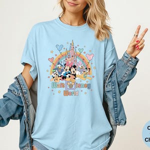Puede incluir: Camiseta azul claro con un gr&aacute;fico colorido de personajes de Disney, un arco&iacute;ris y un castillo. El texto "Walt Disney World" est&aacute; impreso debajo. La camiseta es Comfort Colors Chambray.
