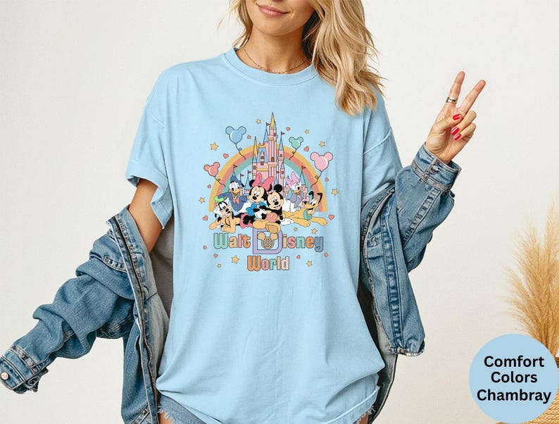 Puede incluir: Camiseta azul claro con un gr&aacute;fico colorido de personajes de Disney, un arco&iacute;ris y un castillo. El texto "Walt Disney World" est&aacute; impreso debajo. La camiseta es Comfort Colors Chambray.