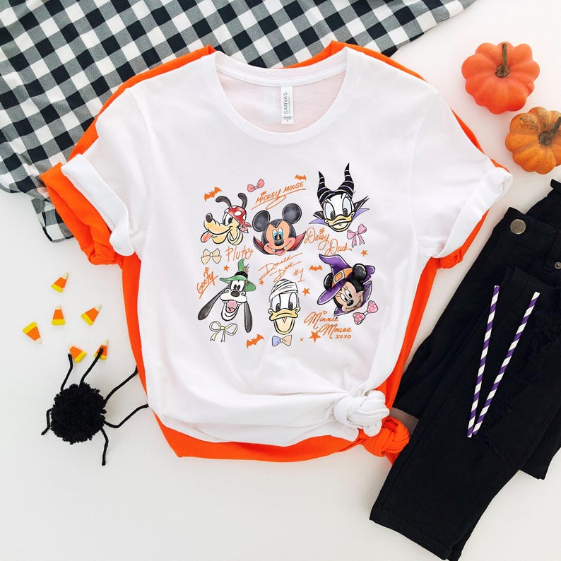 Puede incluir: Camiseta blanca con ribete naranja con retratos de personajes de dibujos animados, incluyendo Mickey Mouse, Minnie Mouse y Donald Duck. La camiseta tiene un tema de Halloween con murci&eacute;lagos y calabazas. Tambi&eacute;n se ven pantalones negros y caramelos de Halloween.