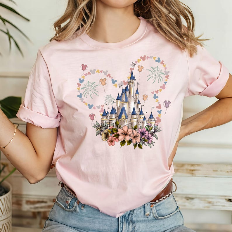 May include: A light pink t-shirt featuring a colorful illustration of a fairytale castle with blue turrets, surrounded by flowers and Mickey Mouse-shaped designs. The design is centered on the shirt, creating a whimsical and cheerful aesthetic.