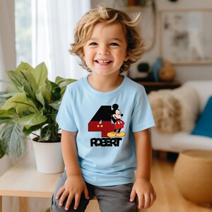 Puede incluir: Una camiseta azul claro con un gr&aacute;fico de Mickey Mouse y el n&uacute;mero 4. El n&uacute;mero 4 es rojo y negro con detalles amarillos. El nombre "ROBERT" est&aacute; impreso debajo en negro.