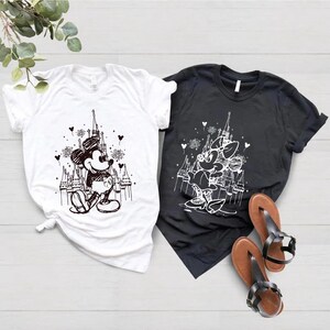 May include: Two t-shirts with a black and white sketch of Mickey Mouse and Minnie Mouse in front of Cinderella's Castle. The t-shirts are white and black.