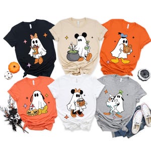 May include: A collection of six Halloween-themed t-shirts. Each shirt features a cartoon ghost character with Mickey Mouse ears and various Halloween props. The shirts are in black, beige, orange, white, and gray.