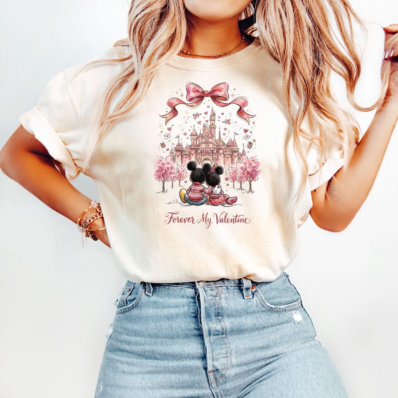 May include: Cream-colored t-shirt featuring a graphic of Mickey and Minnie Mouse embracing in front of a castle, with pink trees and a pink bow. The text "Forever My Valentine" is below the image.