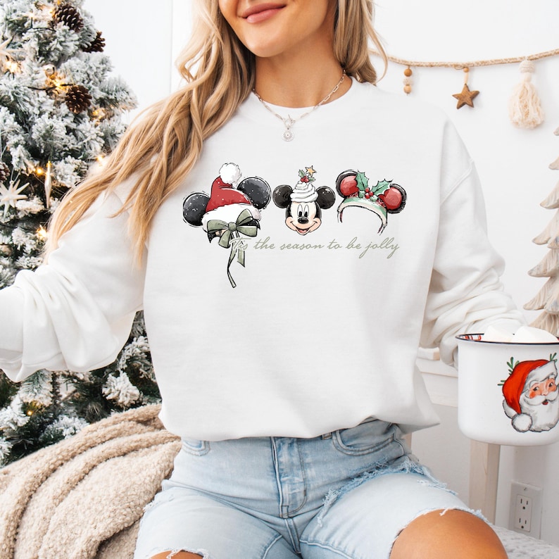 May include: White sweatshirt featuring a festive Mickey Mouse design with Christmas-themed ears and the phrase "the season to be jolly." The graphic includes a Santa hat, holly, and a bow. A white mug with a Santa Claus design is also visible.
