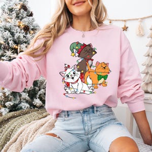 May include: A light pink sweatshirt featuring a graphic of three cartoon cats adorned with Christmas lights and festive accessories. The cats are brown, white, and orange, with a green elf hat and a green bow tie. The sweatshirt is a long-sleeved crew neck.