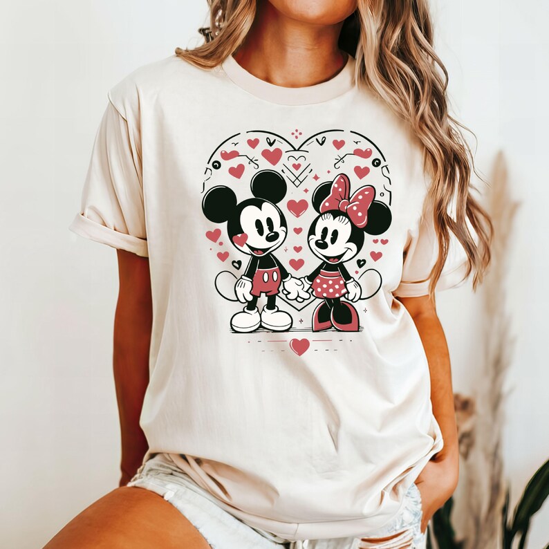 May include: A white t-shirt with a graphic of Mickey Mouse and Minnie Mouse holding hands inside a heart shape. The heart is surrounded by small red hearts and other decorative elements.