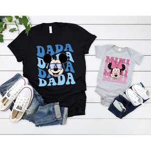 May include: A black t-shirt with a blue and white graphic of Mickey Mouse wearing sunglasses and the word "DADA" repeated. A grey bodysuit with a pink and white graphic of Minnie Mouse and the word "MIMI" repeated.