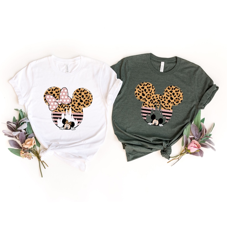 May include: Two t-shirts with a graphic of Mickey Mouse and Minnie Mouse ears with a leopard print pattern. The ears are over a pink and black striped background with a silhouette of the Cinderella castle. The t-shirts are white and green.