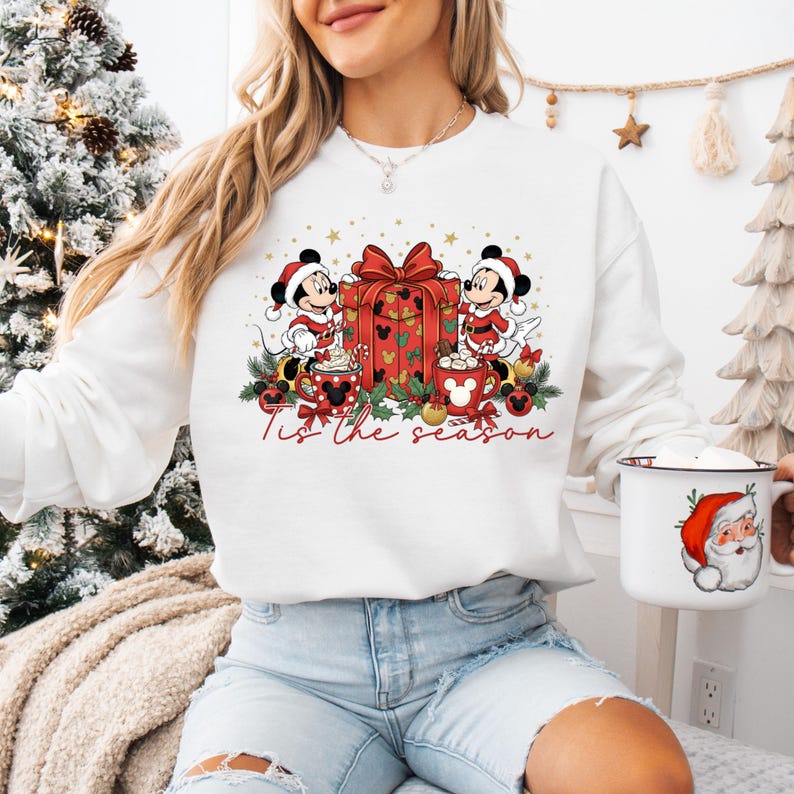 May include: White sweatshirt featuring Mickey and Minnie Mouse in Santa hats, with a red gift box and mugs of hot chocolate. The text "Tis the season" is written in red script. A mug with Santa Claus is held in the hand.