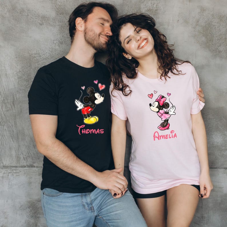 May include: Black t-shirt with a Mickey Mouse graphic and the name "Thomas" in pink. A light pink t-shirt with a Minnie Mouse graphic and the name "Amelia" in pink. Both shirts feature heart designs.
