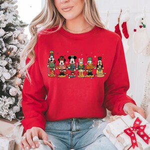 May include: A red crewneck sweatshirt featuring a festive design of Disney characters dressed as nutcrackers. The characters are arranged in a row, with colorful details and holiday lights. The sweatshirt is a solid red color.