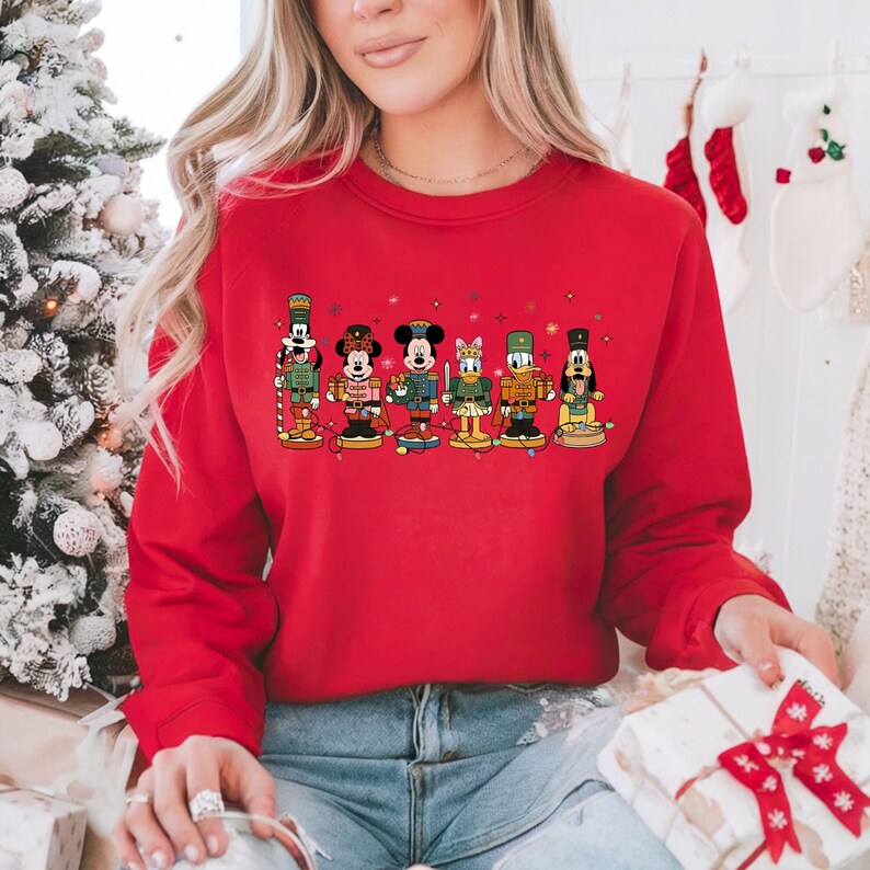 May include: A red crewneck sweatshirt featuring a festive design of Disney characters dressed as nutcrackers. The characters are arranged in a row, with colorful details and holiday lights. The sweatshirt is a solid red color.