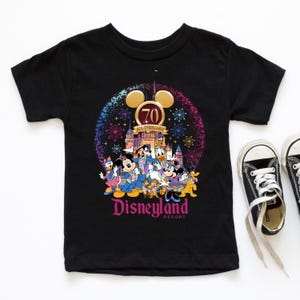 Op de afbeelding: Zwart T-shirt met een kleurrijke afbeelding van Mickey Mouse en zijn vrienden die het 70-jarig jubileum van Disneyland vieren. De afbeelding bevat vuurwerk, het Disneyland-kasteel en de tekst "Disneyland Resort".