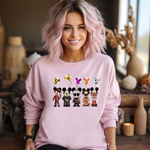 May include: A light pink sweatshirt featuring a cartoon design of Mickey Mouse characters dressed as horror movie villains. Each character holds a balloon with a matching design. The design includes Freddy Krueger, Jason Voorhees, and others.