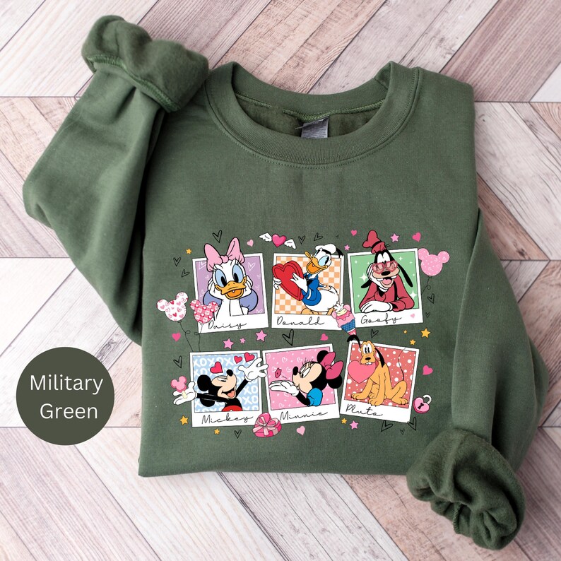 K&ouml;nnte beinhalten: Ein gr&uuml;ner Pullover mit einem Aufdruck von Disney-Charakteren in Polaroid-Rahmen. Die Charaktere sind Mickey Mouse, Minnie Mouse, Pluto, Daisy Duck, Donald Duck und Goofy. Der Druck enth&auml;lt Herzen und die Namen der einzelnen Charaktere.