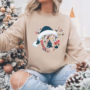 May include: Beige sweatshirt featuring a festive design. The design includes a navy blue Santa hat and a Mickey Mouse head shape filled with Christmas-themed elements like a Christmas tree, presents, and candy canes. The sweatshirt is a neutral color.