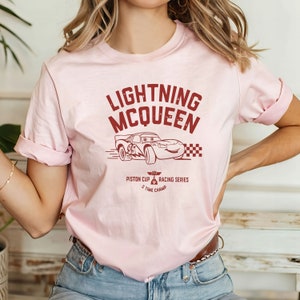 May include: Pink t-shirt with a red graphic of Lightning McQueen from the Cars movie. The graphic includes the text "Lightning McQueen", "Piston Cup", "Racing Series", and "5 Time Champ".