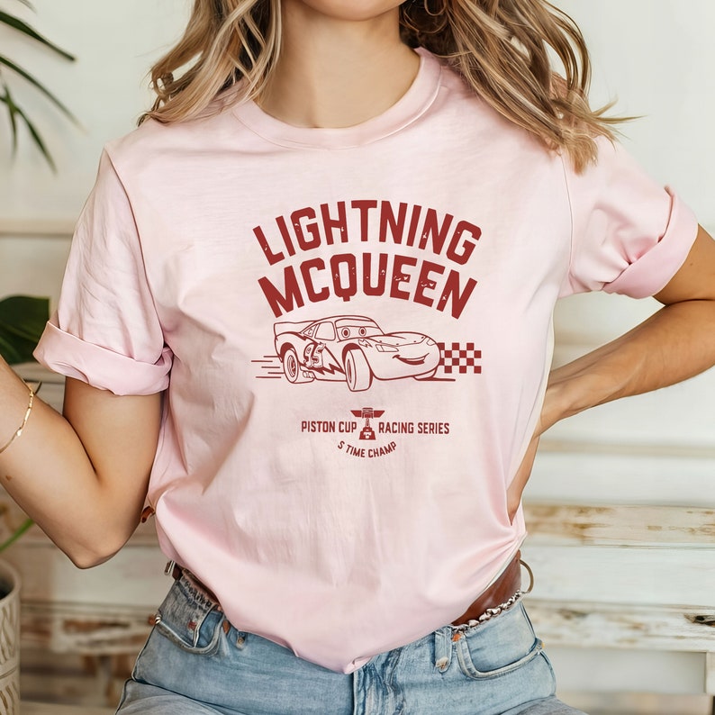 May include: Pink t-shirt with a red graphic of Lightning McQueen from the Cars movie. The graphic includes the text "Lightning McQueen", "Piston Cup", "Racing Series", and "5 Time Champ".