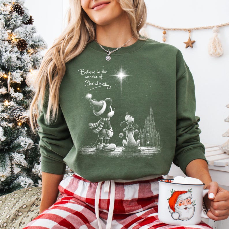 May include: Green crewneck sweatshirt featuring a black and white illustration of Mickey Mouse and Pluto looking at a star, with the text "Believe in the wonder of Christmas." A Santa mug is held in the hand.