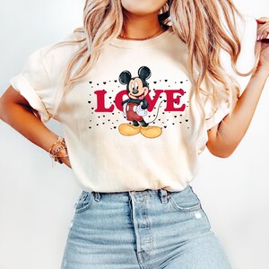 Op de afbeelding: Een wit T-shirt met een rode "LOVE"-afbeelding en een cartoonbeeld van Mickey Mouse. De afbeelding is omringd door kleine zwarte hartjes.