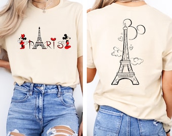 May include: Cream-coloured t-shirt with a front graphic of the Eiffel Tower and the word "PARIS" with Mickey and Minnie Mouse. The back of the shirt shows the Eiffel Tower with Mickey Mouse ears, clouds, and hearts.