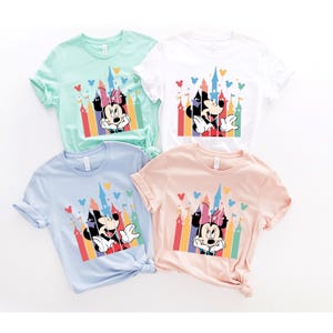 May include: Four t-shirts with different colored backgrounds featuring Mickey Mouse and Minnie Mouse in front of a colorful castle with balloons. The t-shirts are light green, white, light blue, and light pink.