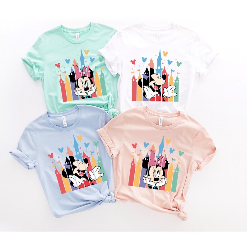 May include: Four t-shirts with different colored backgrounds featuring Mickey Mouse and Minnie Mouse in front of a colorful castle with balloons. The t-shirts are light green, white, light blue, and light pink.
