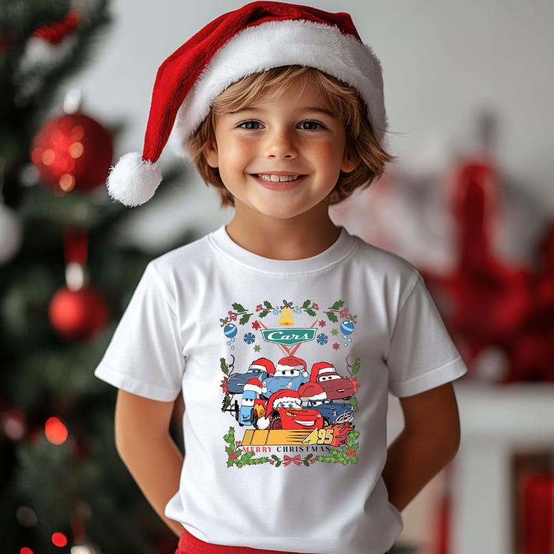 May include: A white t-shirt featuring a Cars-themed Christmas design. The graphic includes several Cars characters wearing Santa hats, the word "Cars", and the text "Merry Christmas". The child is wearing a red and white Santa hat.