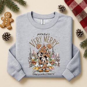 Puede incluir: Sudadera gris claro con un dise&ntilde;o festivo de Mickey Mouse y sus amigos celebrando la Navidad. El dise&ntilde;o incluye el texto "Mickey's Very Merry Christmas Party". Un hombre de jengibre y pi&ntilde;as est&aacute;n en el fondo.