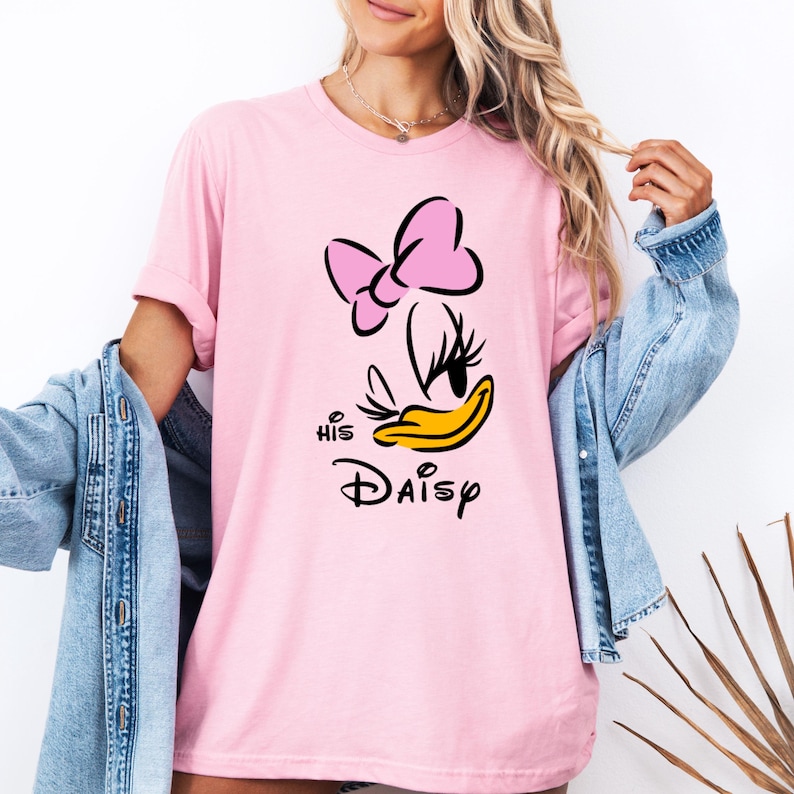 May include: A light pink t-shirt featuring a cartoon Daisy Duck design. The design includes a pink bow, black eyes, and a yellow beak, with the text "His Daisy" in black. The shirt has short sleeves.
