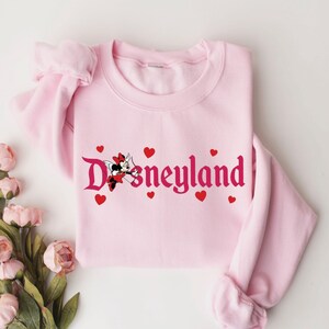 May include: Pink sweatshirt featuring a pink and red graphic of Minnie Mouse with wings and an arrow, and the text "Disneyland" in a pink gothic font.