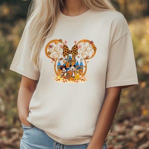 Pode incluir: Camiseta bege com um gr&aacute;fico colorido de Mickey e Minnie Mouse, Pato Donald e Pateta em frente a um castelo. O design inclui um la&ccedil;o de bolinhas, folhas de outono e uma ab&oacute;bora. A camiseta tem mangas curtas e gola redonda.