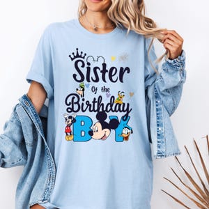 May include: Light blue t-shirt with a graphic of Mickey Mouse and other Disney characters. The text on the shirt reads "Sister of the Birthday Boy".