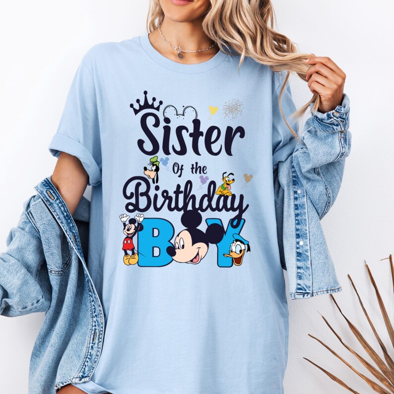 May include: Light blue t-shirt with a graphic of Mickey Mouse and other Disney characters. The text on the shirt reads "Sister of the Birthday Boy".