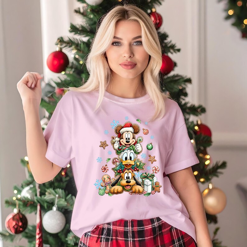 Peut inclure: T-shirt rose clair avec un motif de No&euml;l repr&eacute;sentant Mickey Mouse, Donald Duck et Pluto. Le motif comprend des flocons de neige, des cannes en sucre et des bonhommes en pain d'&eacute;pices. La personne porte le t-shirt devant un sapin de No&euml;l.