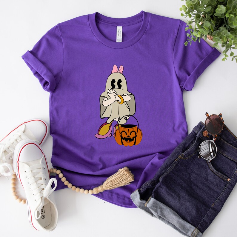 Puede incluir: Camiseta morada con un fantasma de dibujos animados que lleva un lazo rosa y sostiene un cubo de calabaza. El fantasma lleva una pulsera dorada y tiene una expresi&oacute;n de sorpresa.