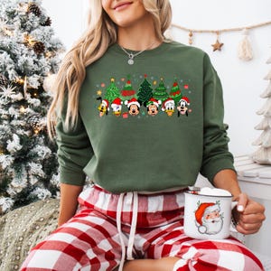 May include: Green sweatshirt featuring a festive design with cartoon Christmas trees and character faces. The characters are wearing Santa hats. The sweatshirt is paired with red and white plaid pajama pants. A white mug with a Santa Claus graphic is held in the person's hand.