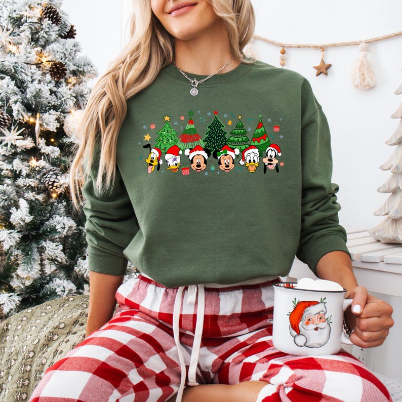 May include: Green sweatshirt featuring a festive design with cartoon Christmas trees and character faces. The characters are wearing Santa hats. The sweatshirt is paired with red and white plaid pajama pants. A white mug with a Santa Claus graphic is held in the person's hand.